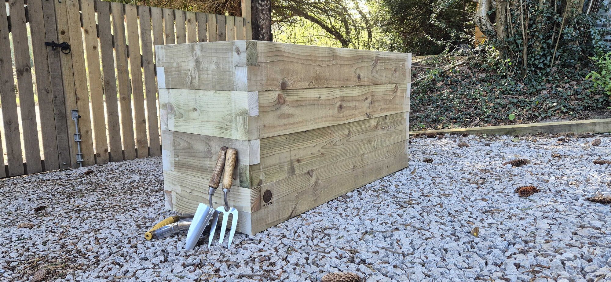 HEWN stacked-timber planter on gravel in a North Wales garden, beside gardening tools